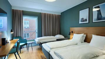 Habitación de hotel con dos camas, un escritorio con silla, una mesita y vista a un edificio de ladrillo a través de una ventana.