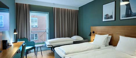 Habitación de hotel con dos camas, un escritorio con silla, una mesita y vista a un edificio de ladrillo a través de una ventana.