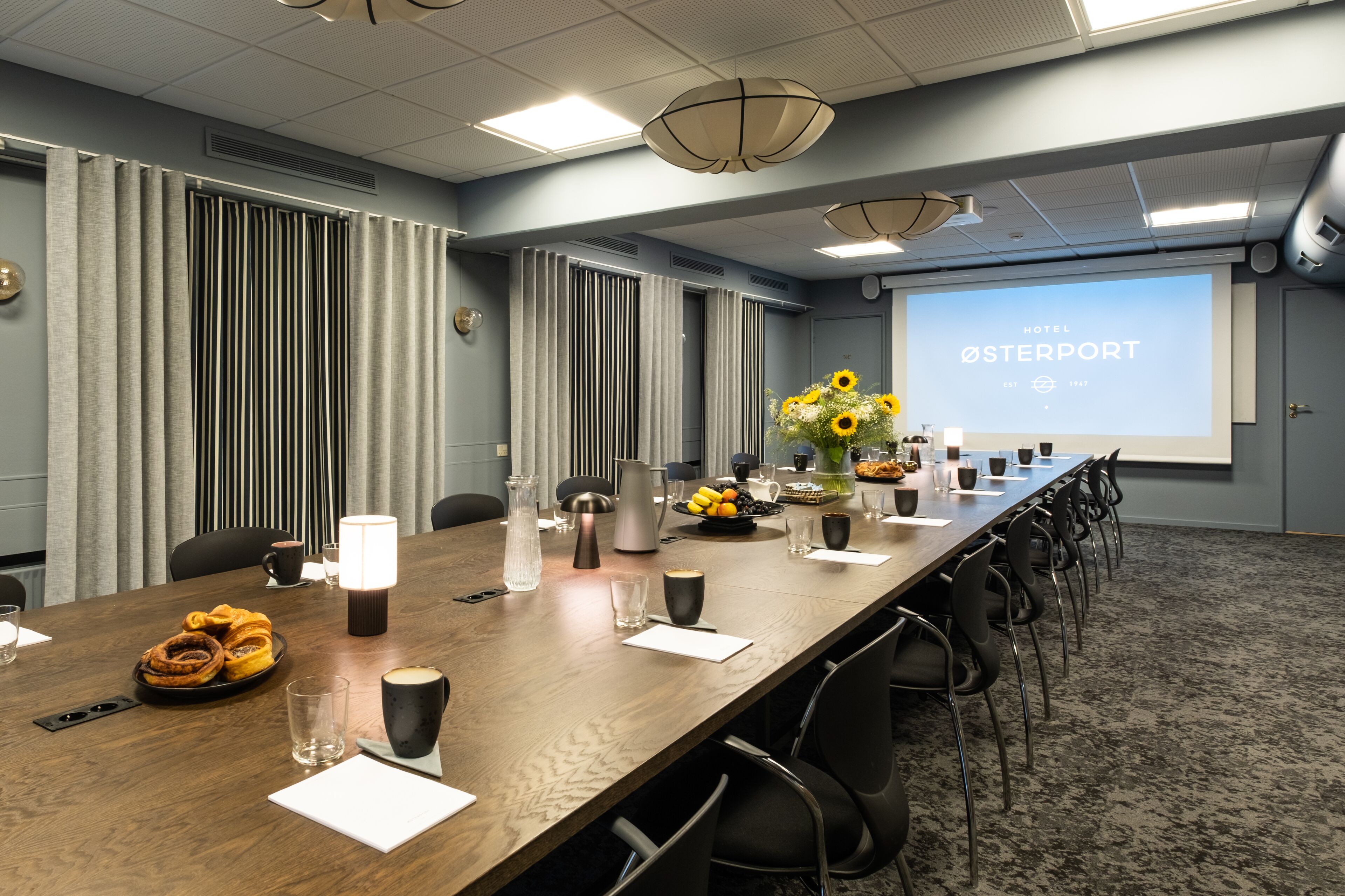 Un salón de conferencias con una larga mesa, sillas, una pantalla de proyector mostrando 'HOTEL ØSTERPORT' y un arreglo floral.