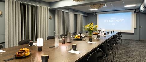 Un salón de conferencias con una larga mesa, sillas, una pantalla de proyector mostrando 'HOTEL ØSTERPORT' y un arreglo floral.
