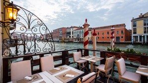 An outdoor dining area with tables and chairs, a railing, and a view of the water and buildings in the background. - CHC Continental, BW Premier Collection (Venice)