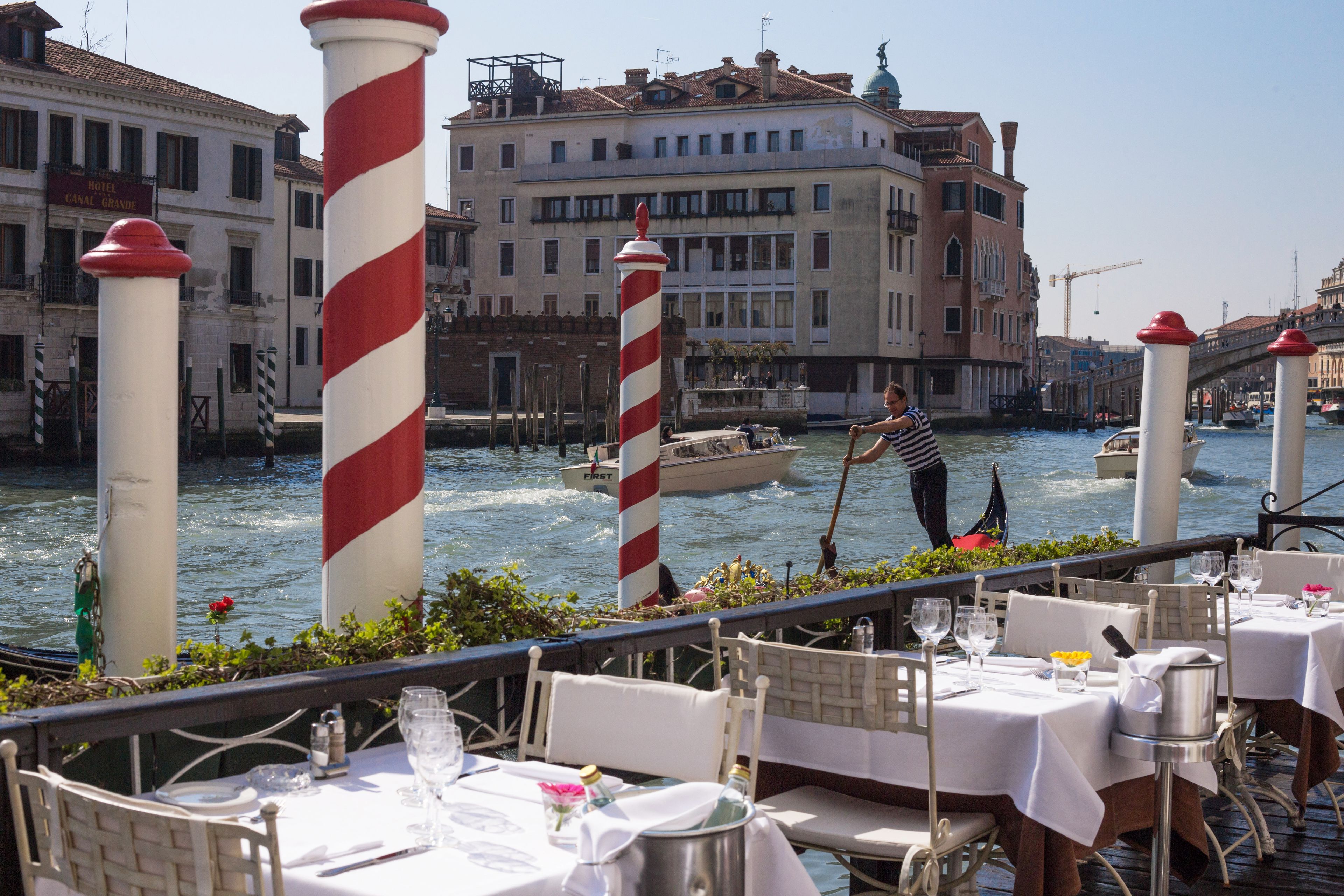 Un restaurant al costat del port amb taules preparades per dinar, un canal amb barques i edificis històrics al fons.