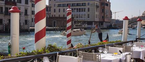 Un restaurant al costat del port amb taules preparades per dinar, un canal amb barques i edificis històrics al fons.