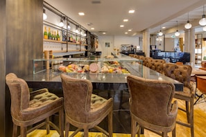 A bar area with a glass counter displaying various food items and bottles. There are leather bar stools and a seating area with cushioned chairs.