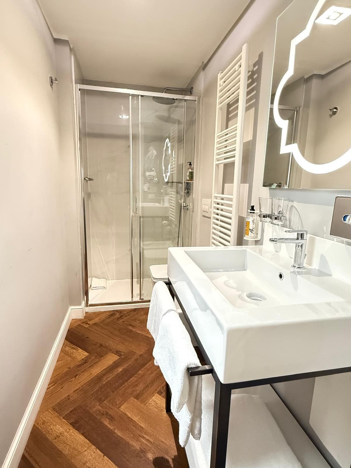 A modern bathroom with a glass-enclosed shower, a white sink with a mirror, and wooden flooring.