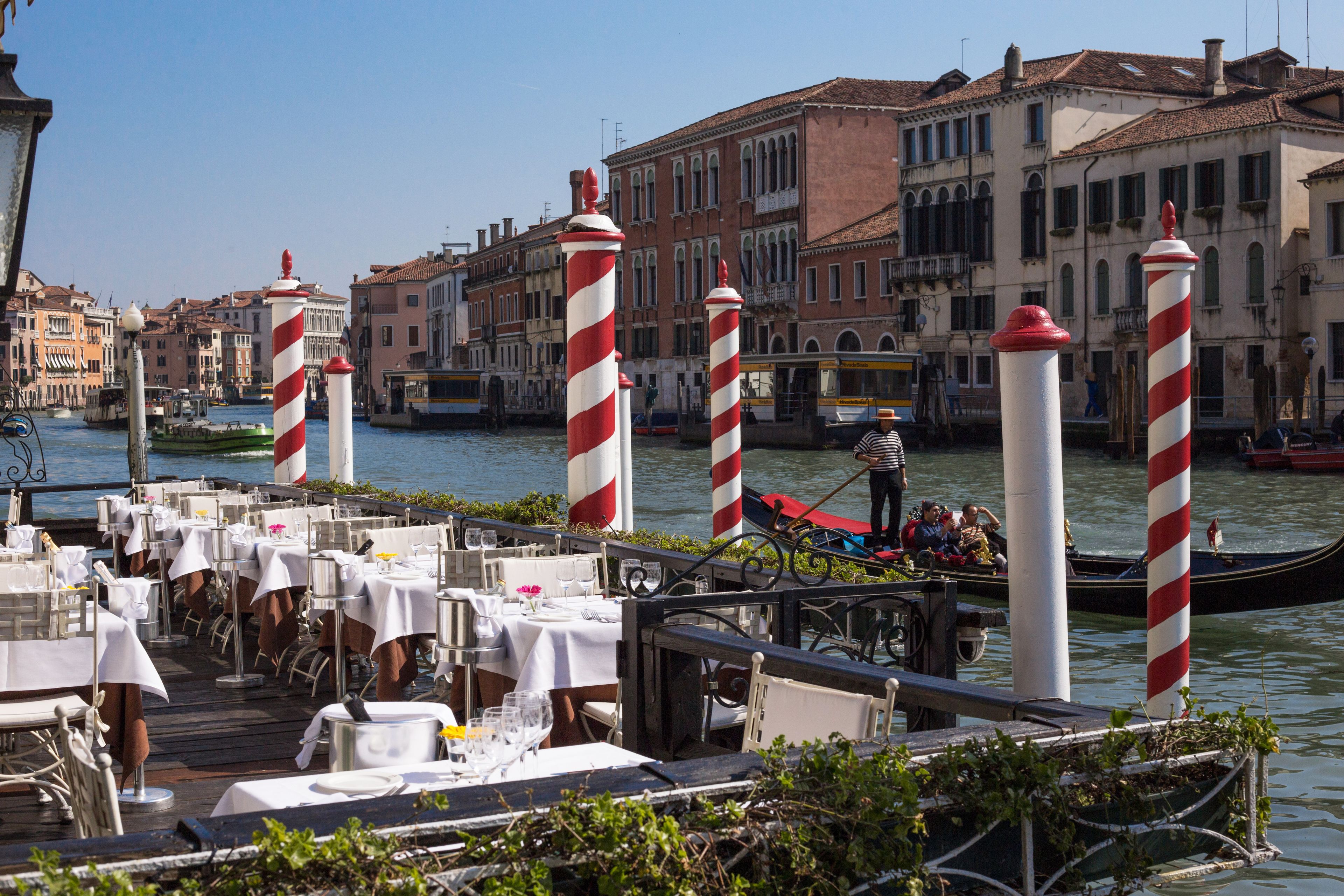 A riverside dining area with tables set for guests, a gondola in the background, and traditional Venetian buildings.