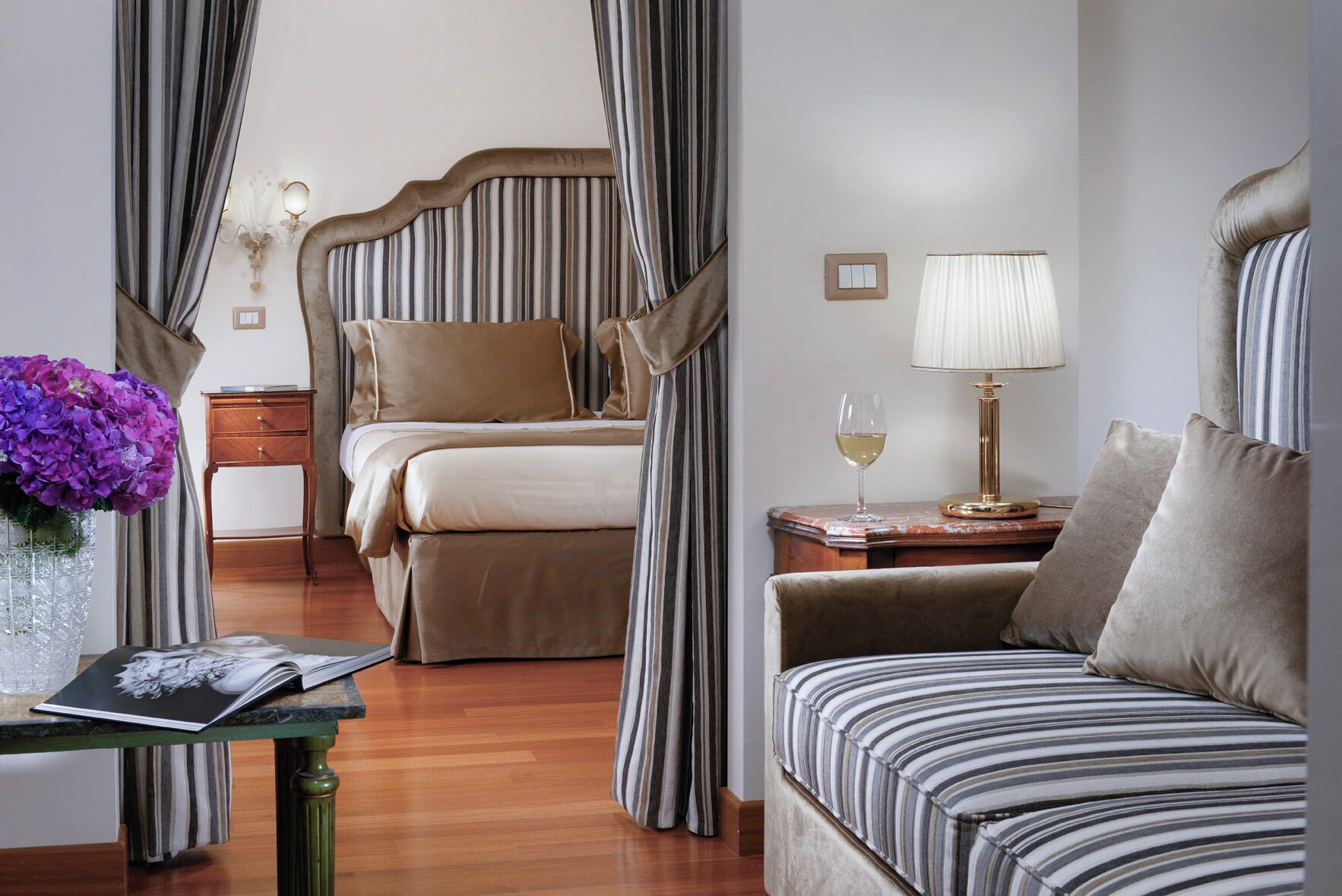 A hotel room with a bed, a sofa, a nightstand with a lamp, a vase with flowers, and a window with curtains.