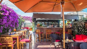 Un área de bar al aire libre con mesas y sillas de madera, una barra y plantas con flores moradas.