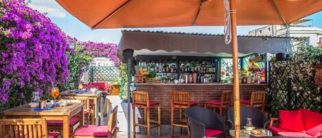An outdoor bar area with wooden tables and chairs, a bar counter, and purple flowering plants.