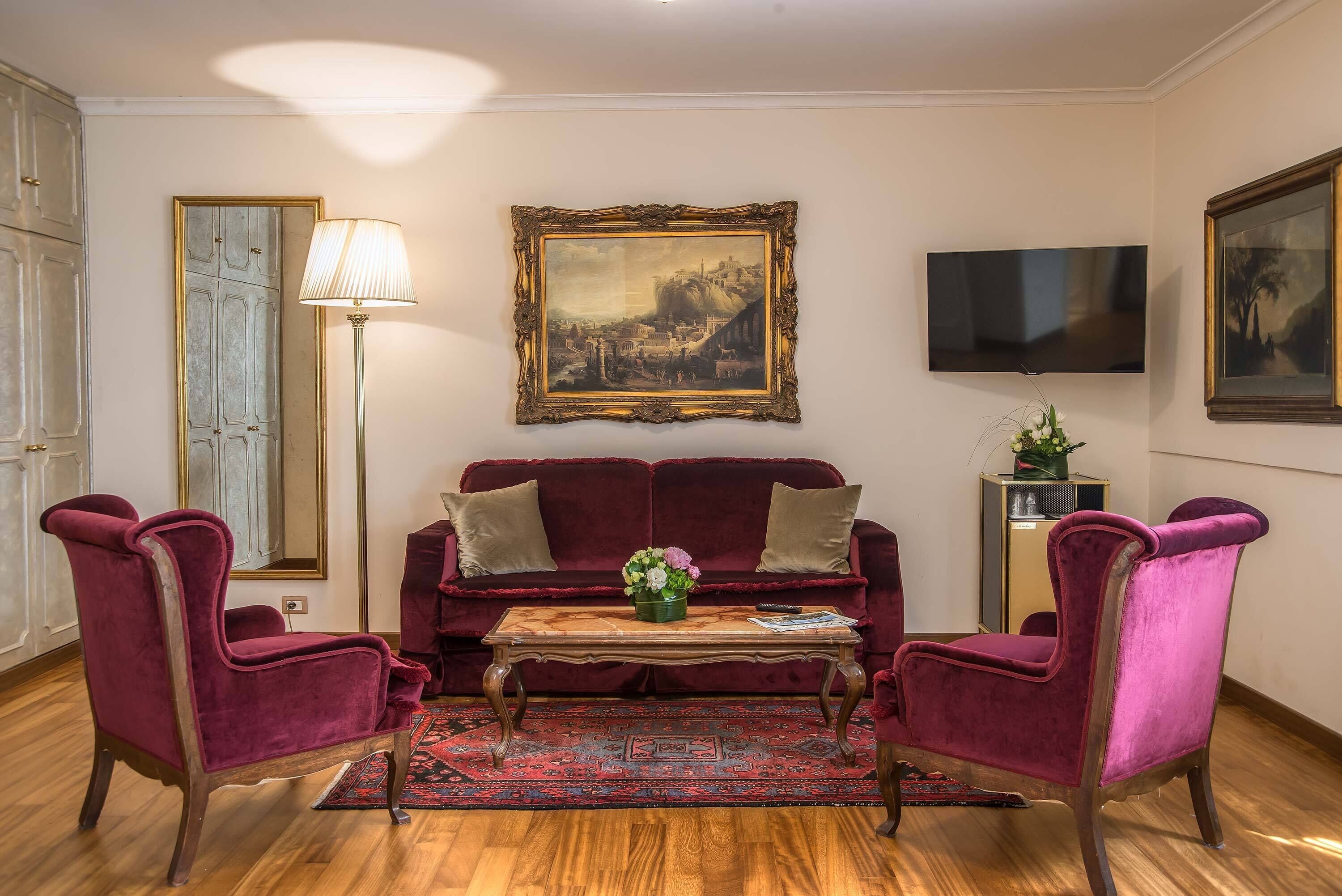 A living room with a red velvet sofa, two matching armchairs, a wooden coffee table, a floor lamp, a framed painting, and a television.