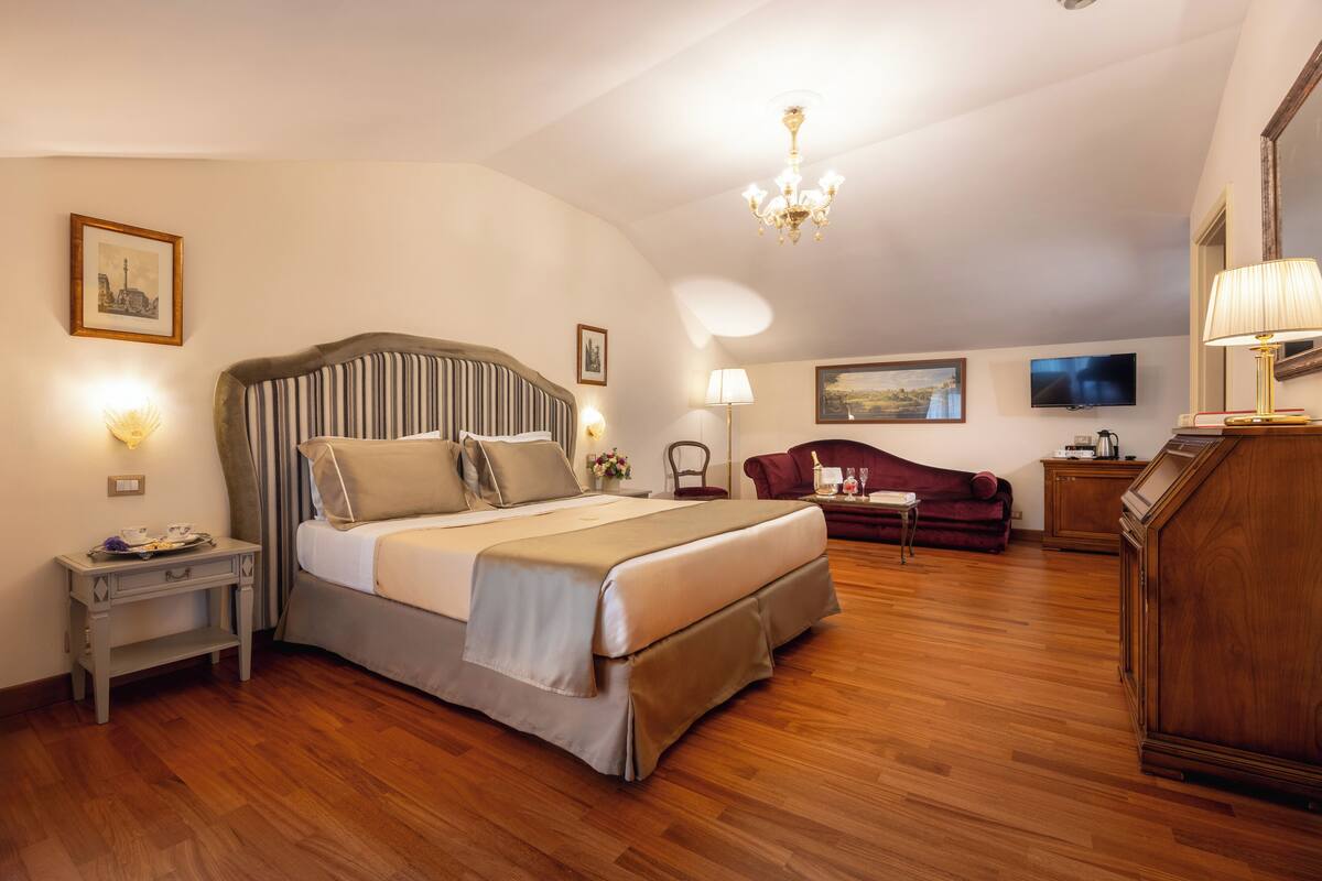A hotel room with a large bed, a wooden floor, a desk, a chair, a TV, and a chandelier.