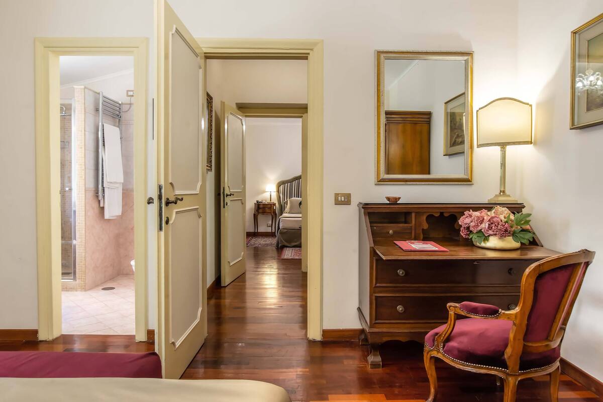 A hotel room with a wooden floor, a bed, a desk with a lamp, a chair, a dresser, and a mirror.