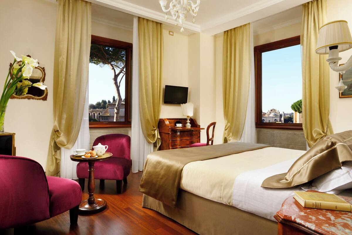A hotel room with a large bed, a desk with a chair, a TV, and a view of a cityscape through the windows.