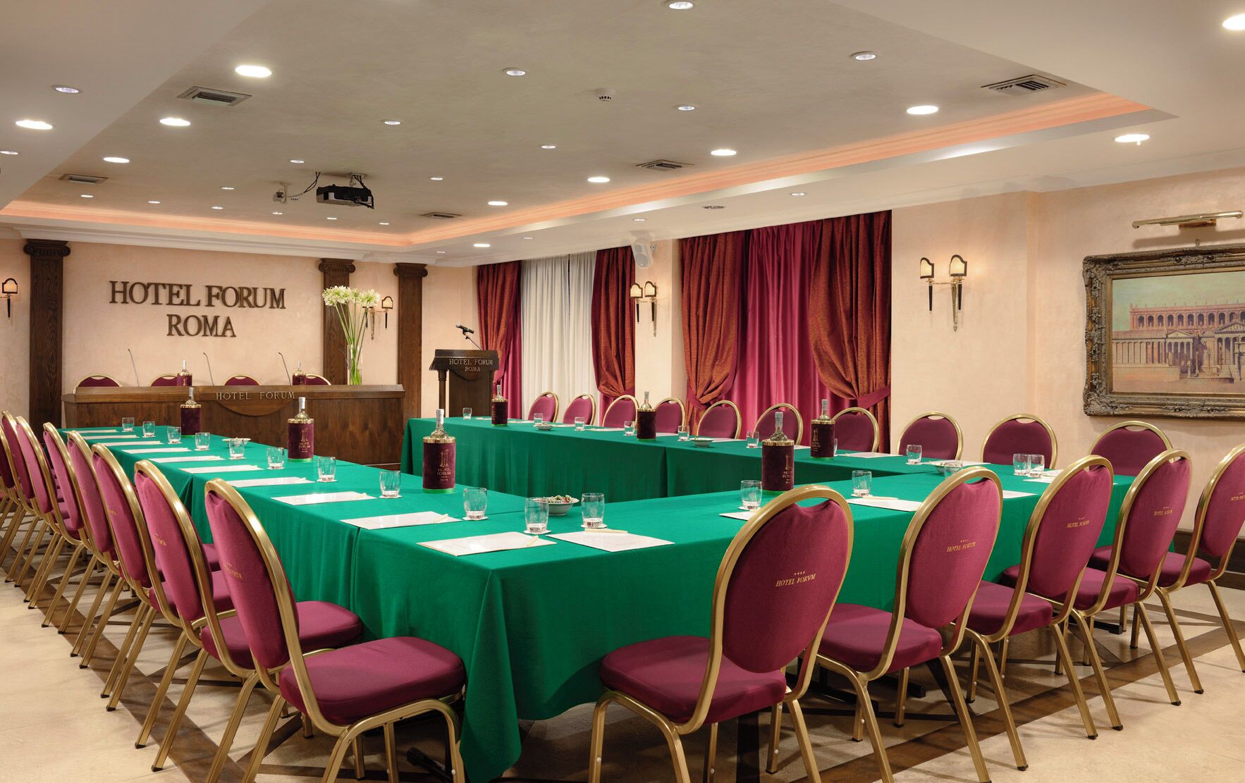 A conference room with green tables and maroon chairs, a podium, and a sign reading 'HOTEL FORUM ROMA'.