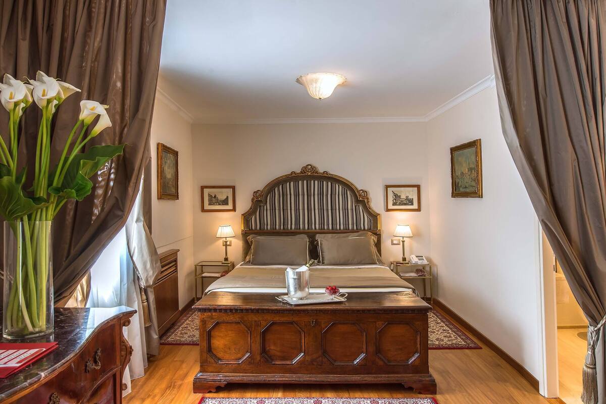A hotel room with a large bed, a wooden dresser, a vase of flowers, and a painting on the wall.