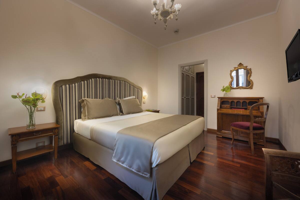 A bedroom with a bed, bedside tables, a desk, a chair, a mirror, and a chandelier.