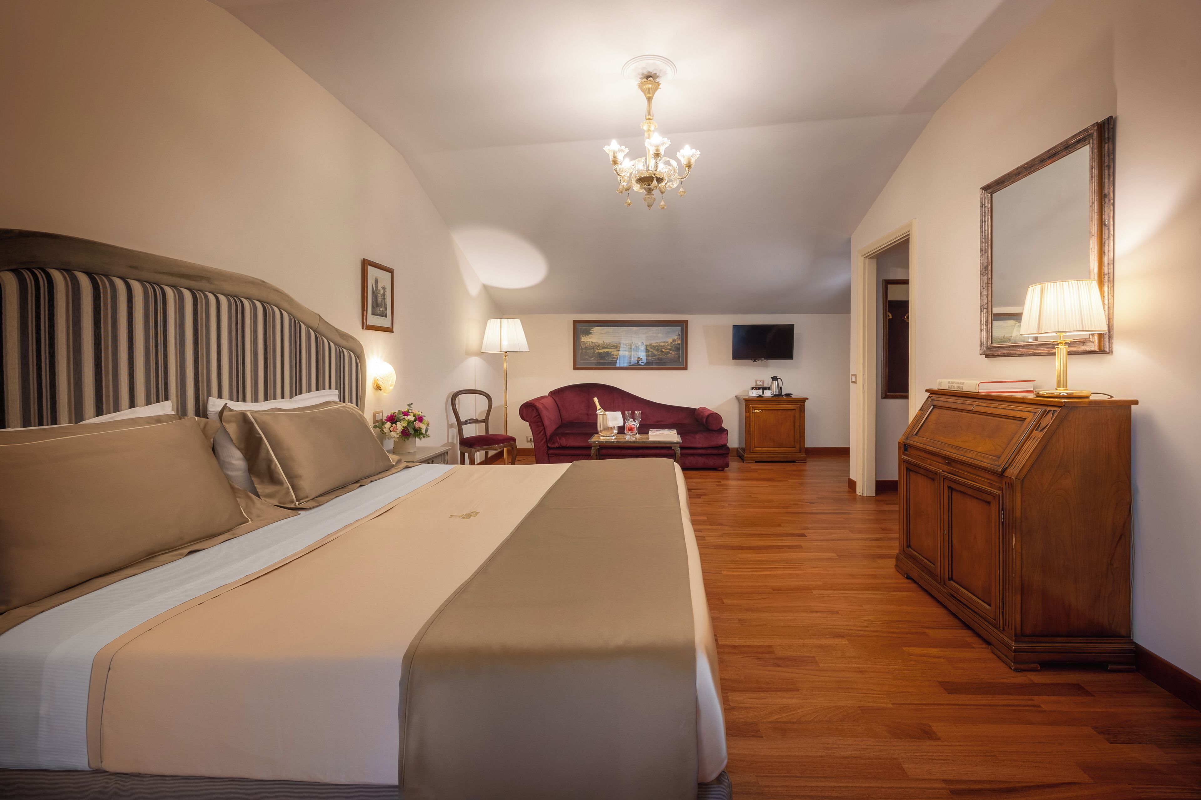Une chambre d’hôtel avec un grand lit, une commode en bois, un fauteuil rouge, une télévision et un lustre.