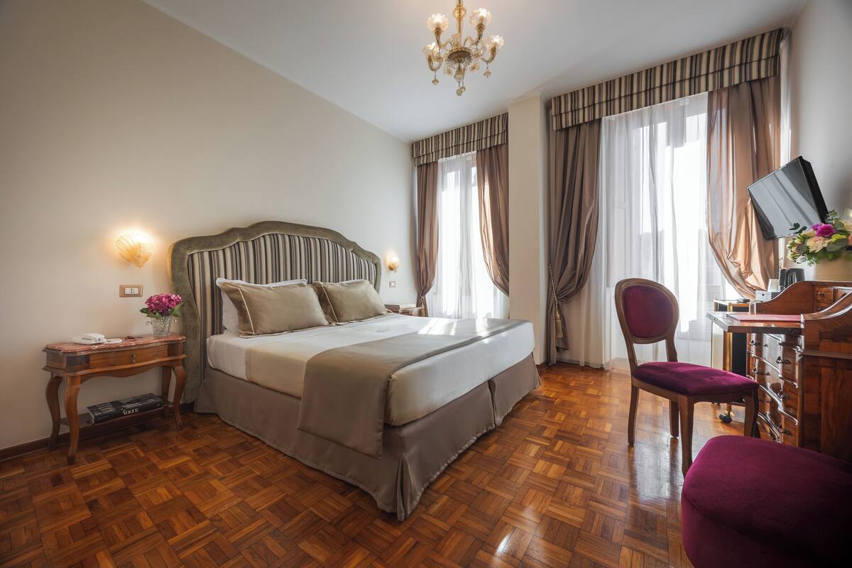 A hotel room with a large bed, a desk with a chair, a chandelier, and a large window with curtains.