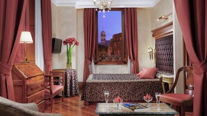 Habitación de hotel con cama, escritorio, silla, mesa con un libro y gafas, florero con flores, y vista de un edificio a través de una ventana.