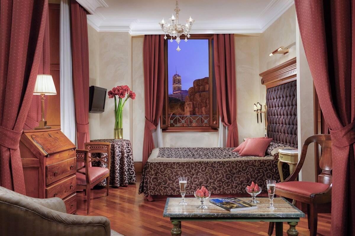 A hotel room with a bed, a desk, a chair, a table with a book and glasses, a vase with flowers, and a view of a building through a window.