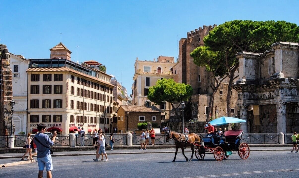 A historic building with a prominent tower, a horse-drawn carriage, and a hotel named 'HOTEL FORUM' in the foreground.