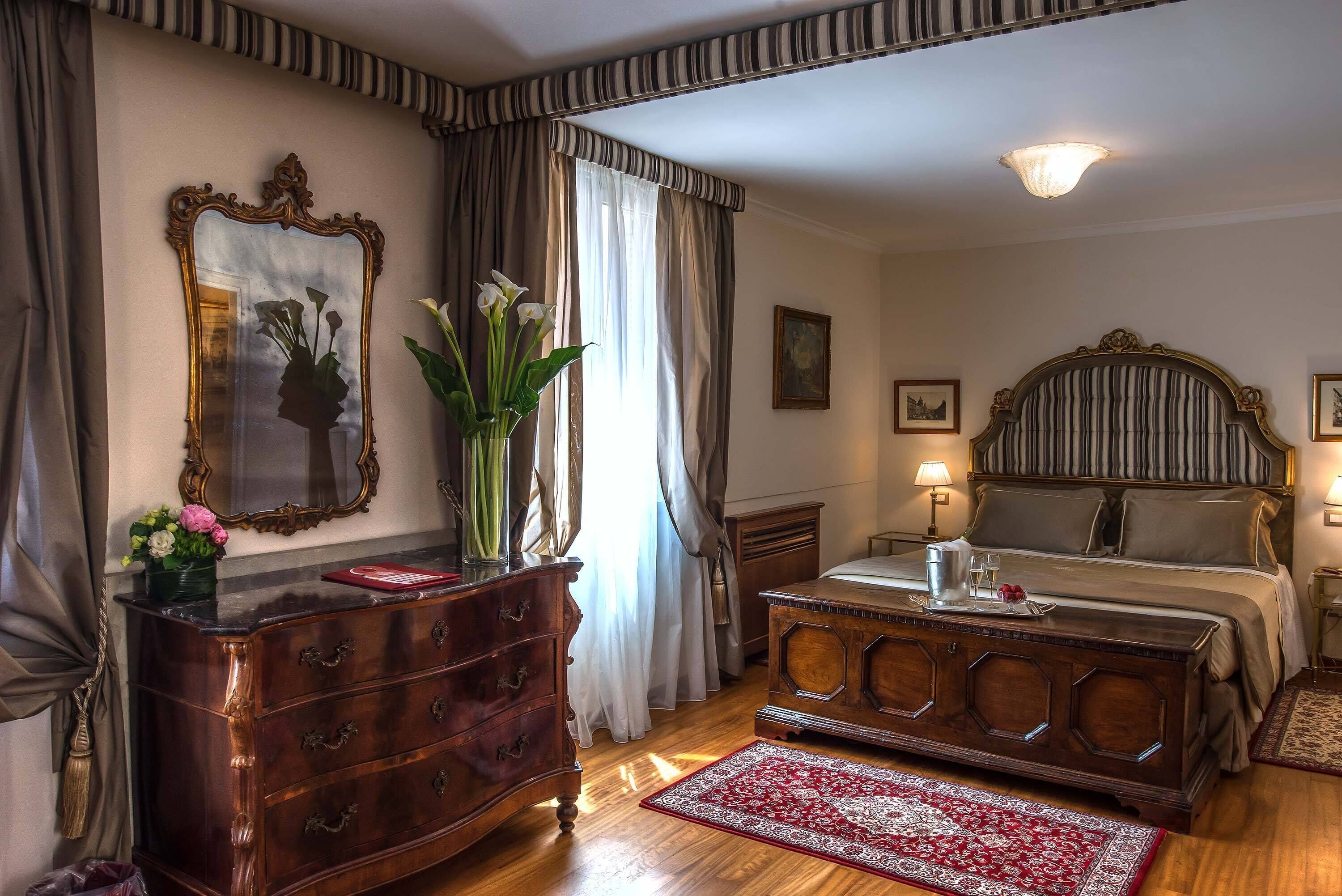 Habitación de hotel con tocador de madera, cama con cabecero, espejo y un florero con flores.