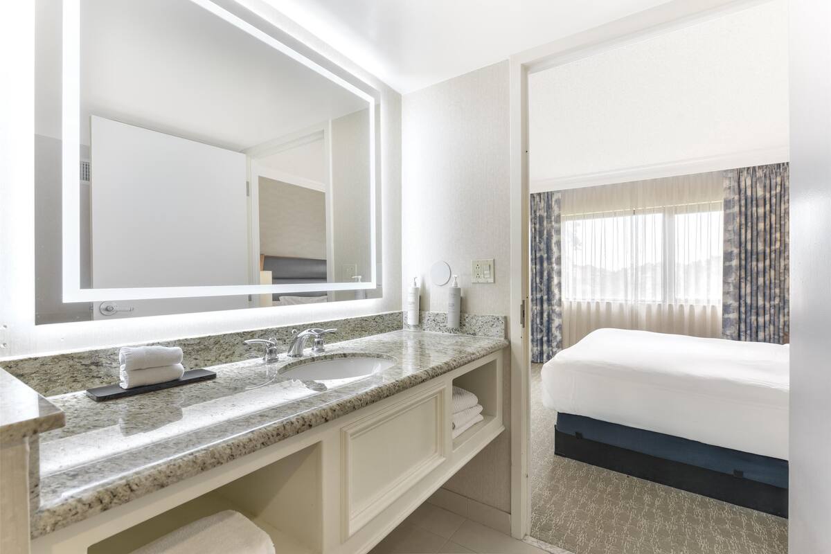 A hotel bathroom with a large mirror, two sinks, a vanity, and a bed with a window view.