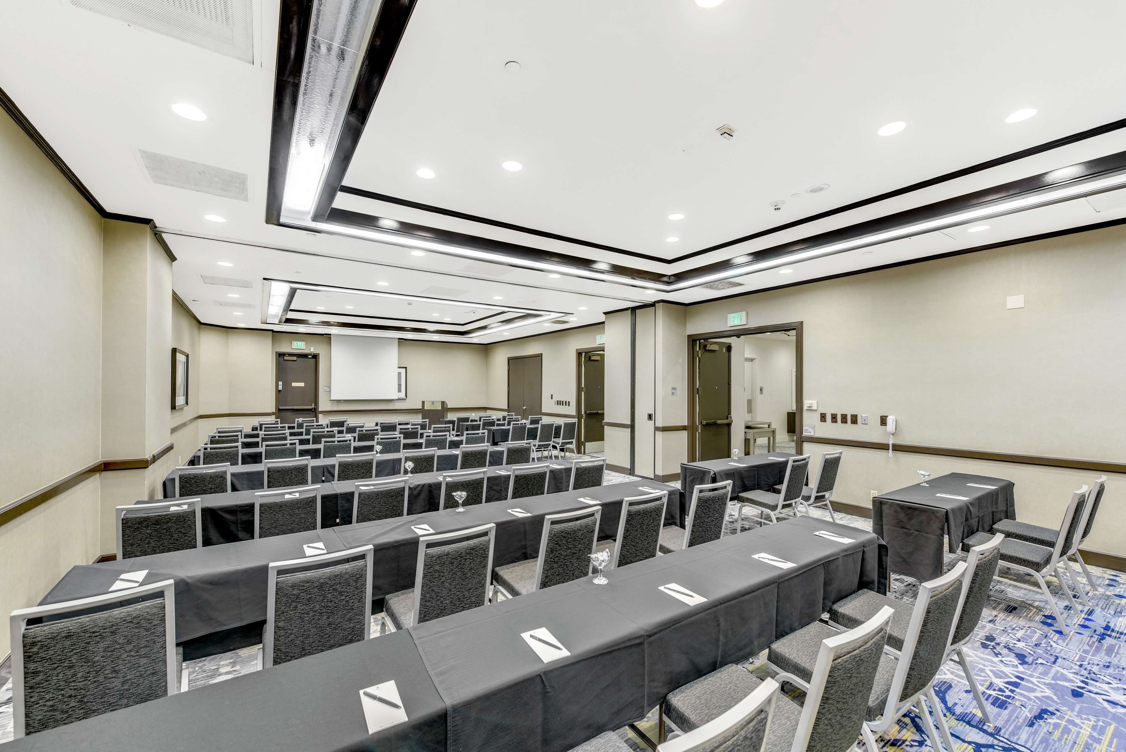Un salón de conferencias con filas de mesas y sillas, equipado con proyectores y pantallas para presentaciones.
