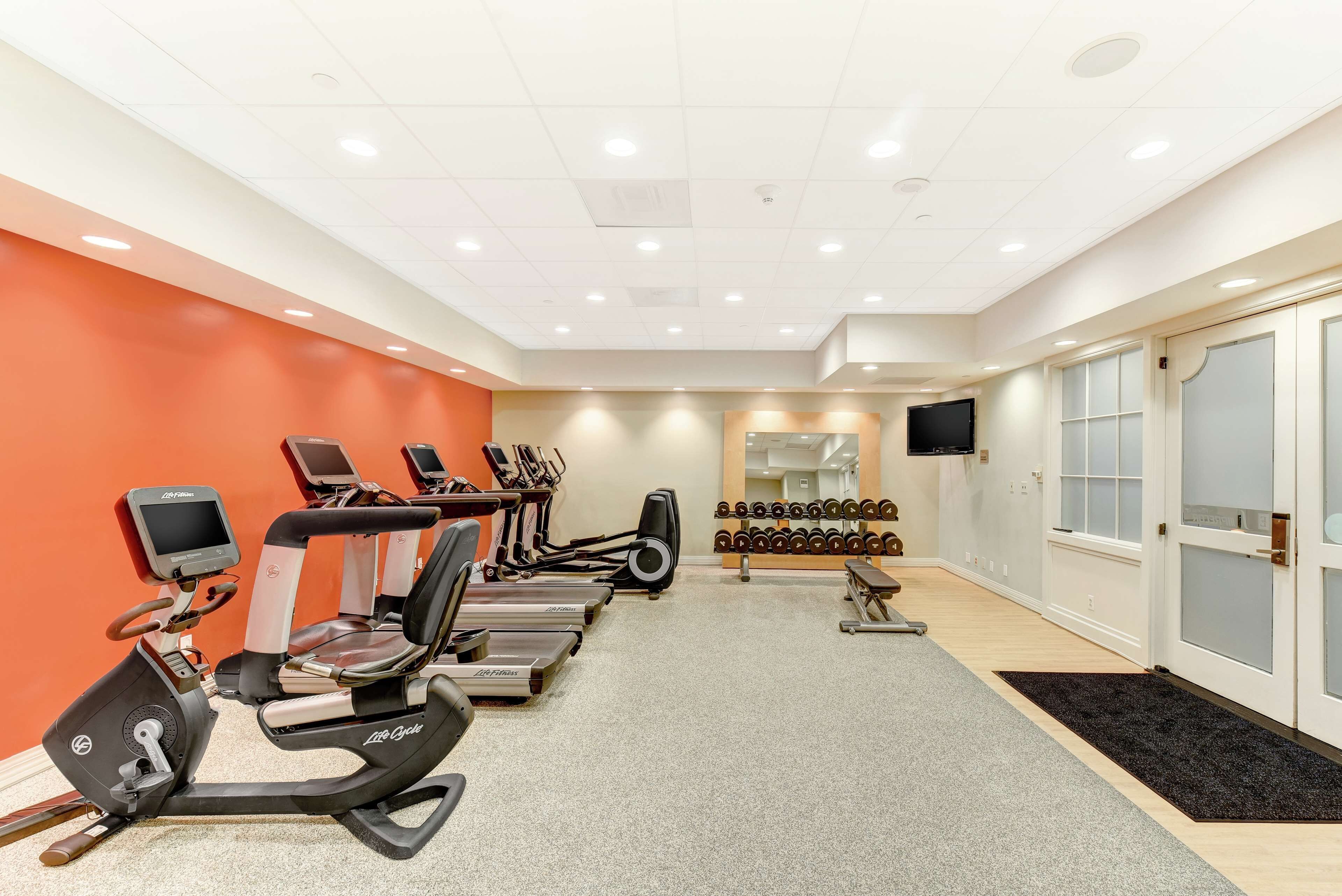 Un gimnasio con máquinas de cardio, mancuernas y un televisor en la pared.