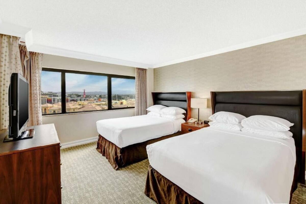 A hotel room with two beds, a TV, a desk, and a large window with a city view.