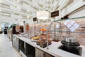 A modern hotel lobby with a high ceiling, a central serving area with food and beverage options, and a sign indicating the location of the oatmeal station.
