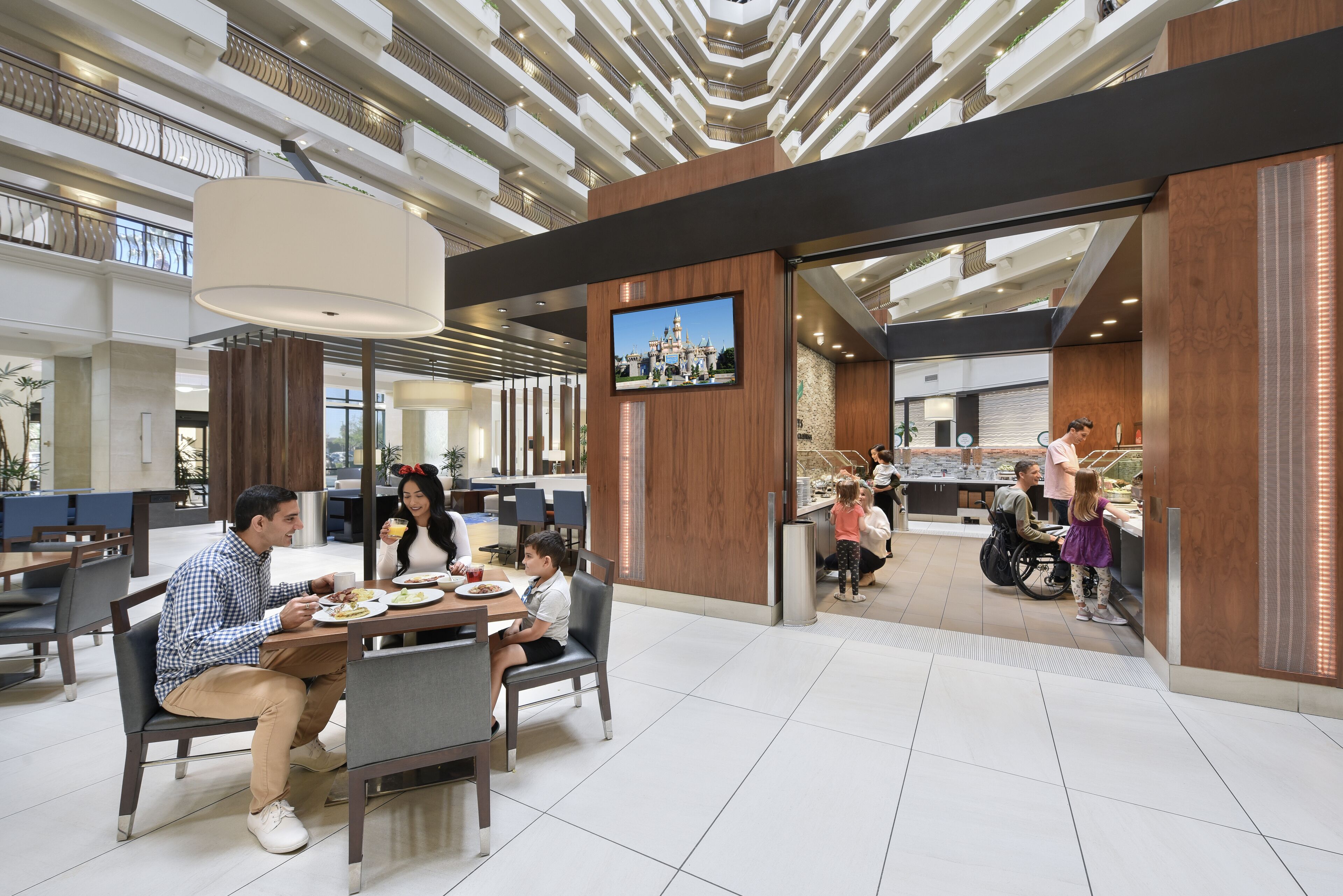 A modern indoor dining area with multiple tables and chairs, a large TV screen, and a view of a building through glass doors.