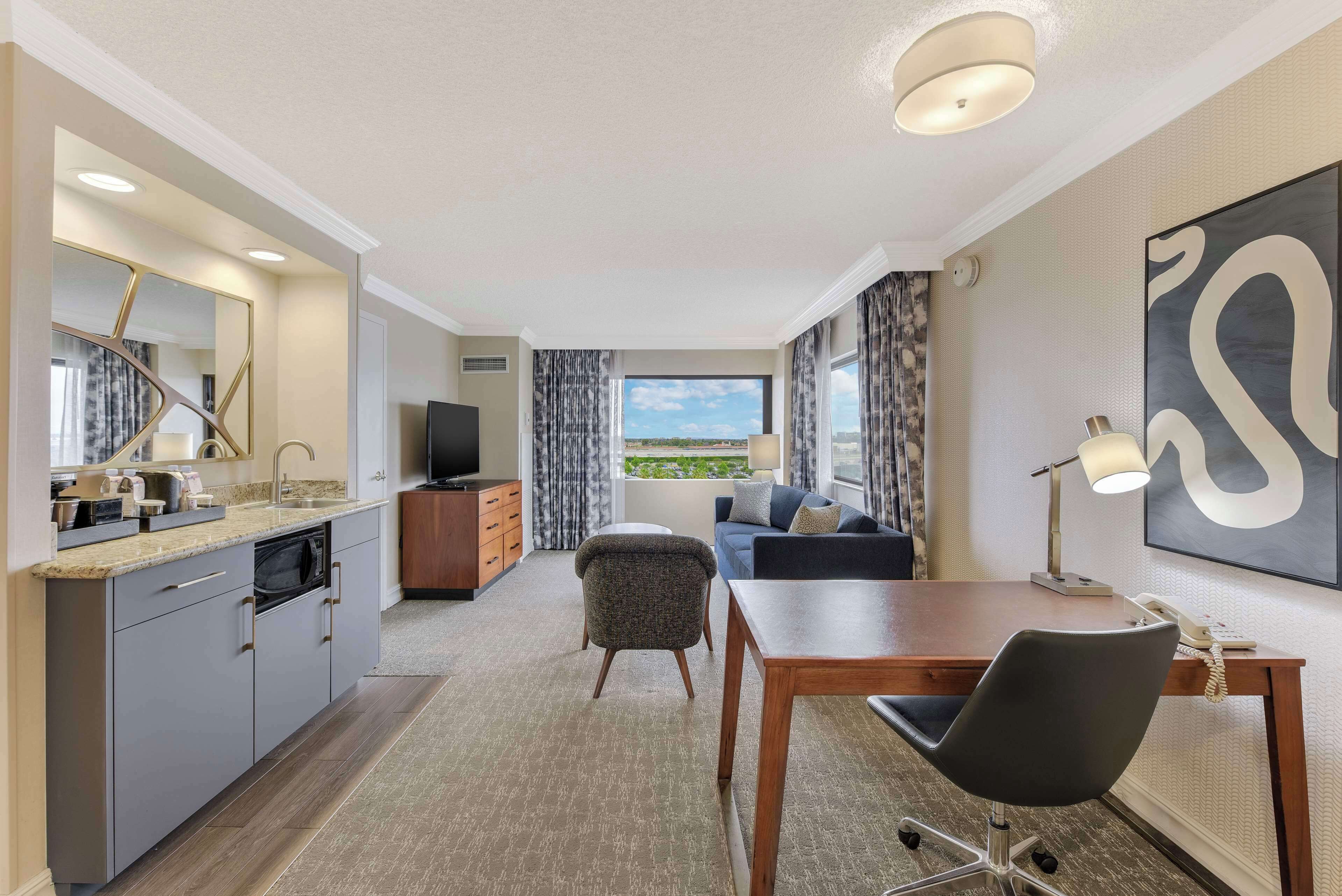 A modern hotel room with a kitchen, a dining area, a sofa, and a large window offering a view.