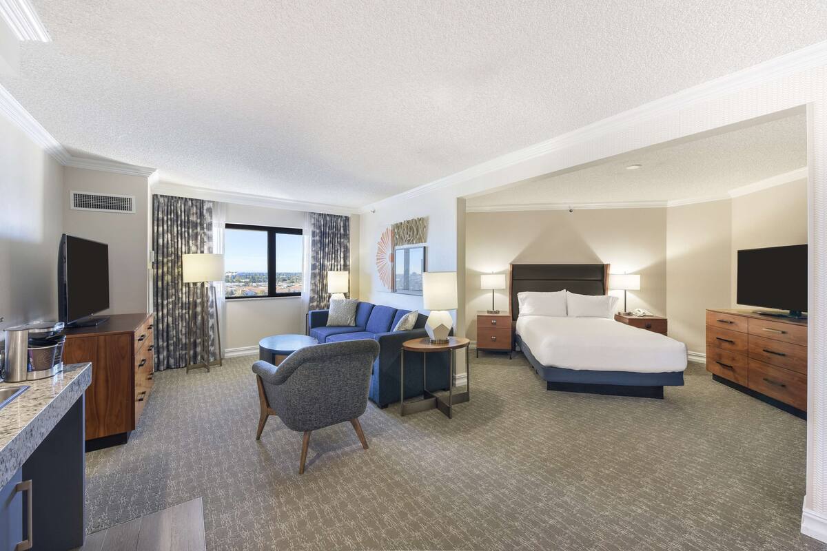 A hotel room with a bed, a desk, a chair, a TV, and a view of the city.