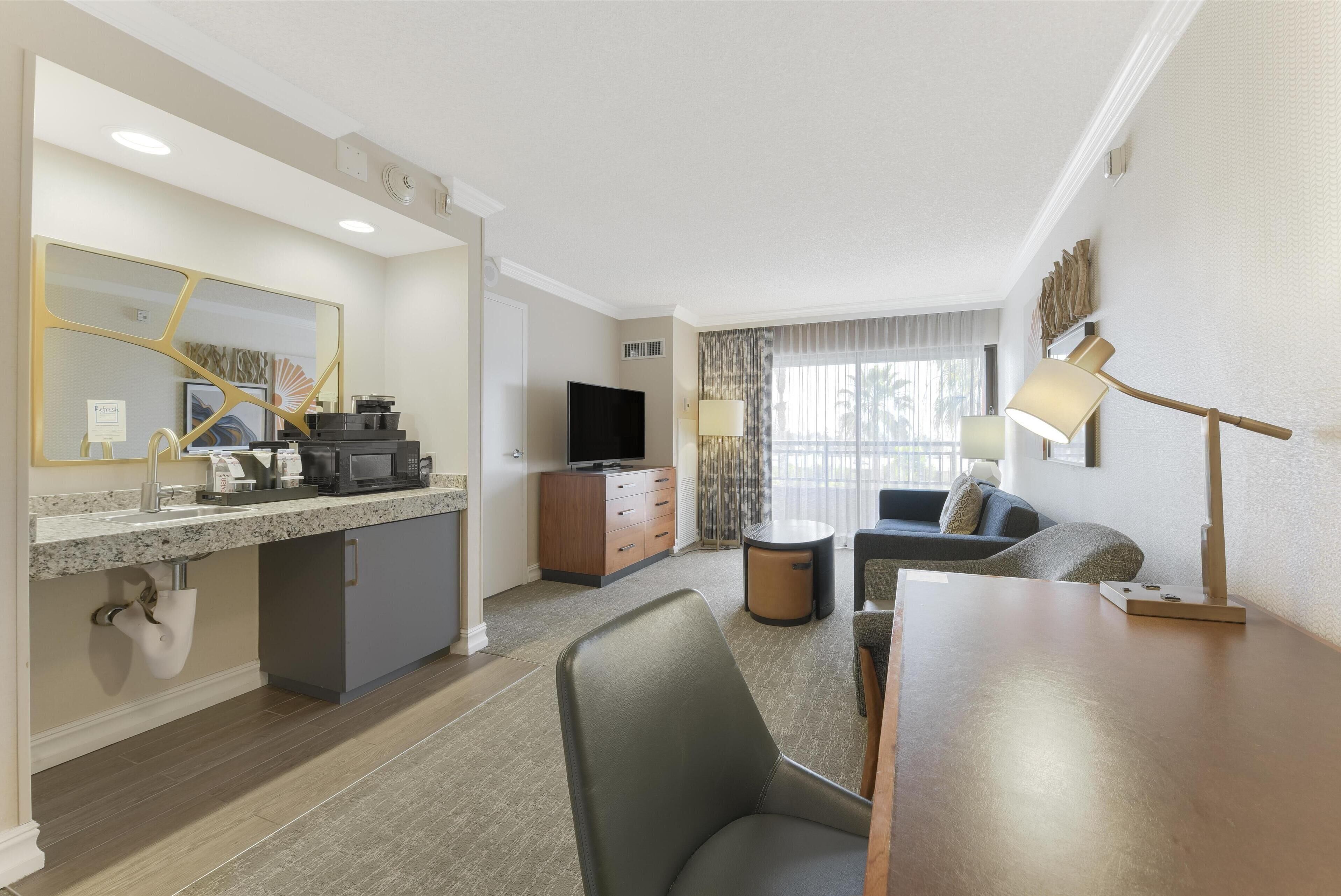 A modern hotel room with a kitchenette, a flat-screen TV, a sofa, and a dining table.