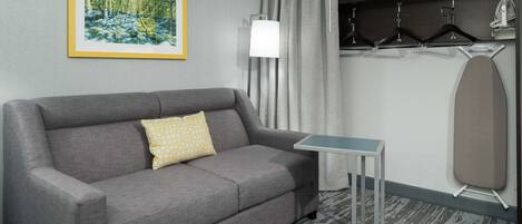 Une chambre d’hôtel avec un canapé gris, une petite table, une lampe, un porte-manteau et un tableau encadré au mur.