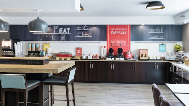 A modern kitchen with dark wood cabinets, a granite countertop, and a central island with seating. There are various kitchen appliances and a chalkboard menu on the wall. - Hampton Inn Los Angeles/Santa Clarita (Stevenson Ranch)