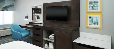 Una habitación de hotel moderna con un televisor de pantalla plana grande montado en un mueble de madera oscura, una silla azul, un escritorio blanco y dos cuadros en la pared.