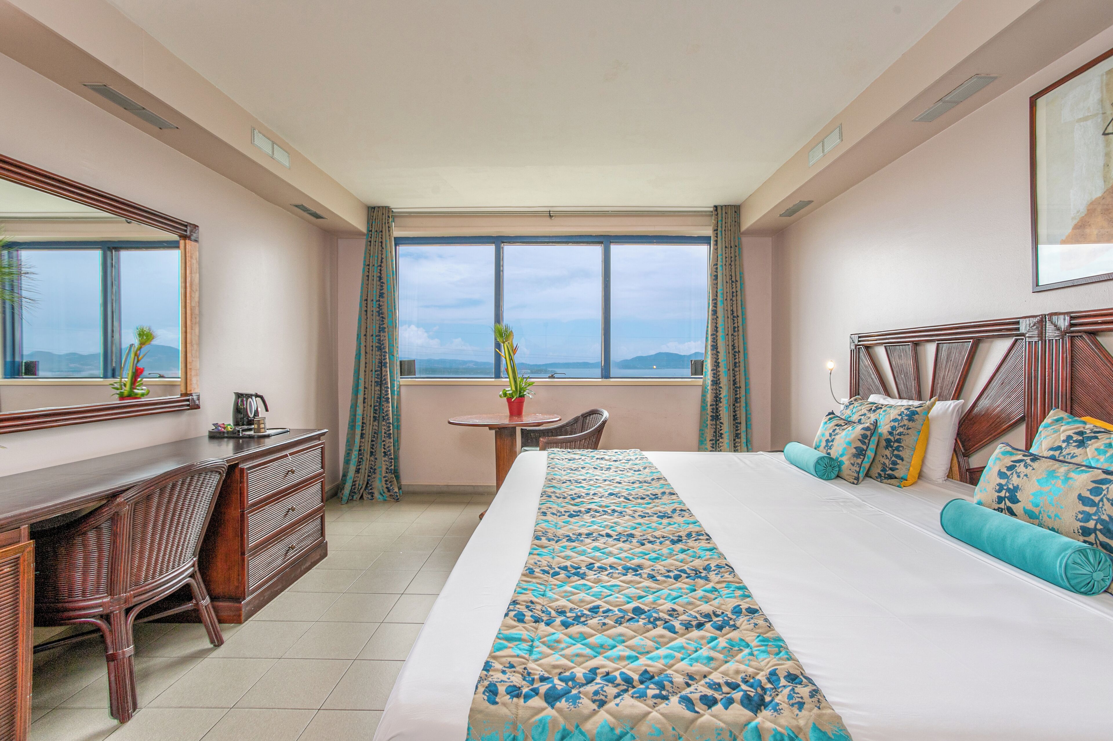 A hotel room with a large bed, a desk, a chair, a small table, and a view of the mountains through the window.