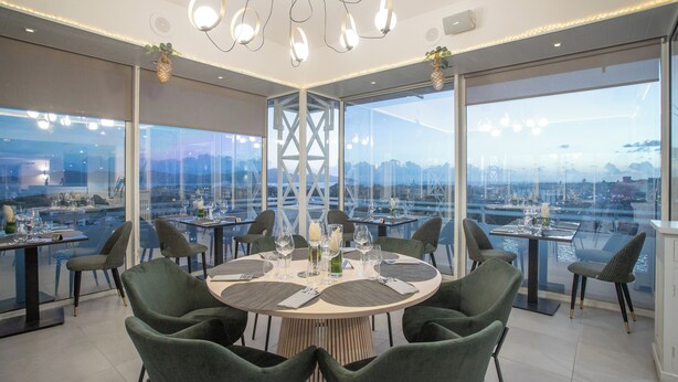 A modern dining area with round tables, green upholstered chairs, and large windows offering a city view. - Karibea Valmenière Hôtel (Fort-de-France)