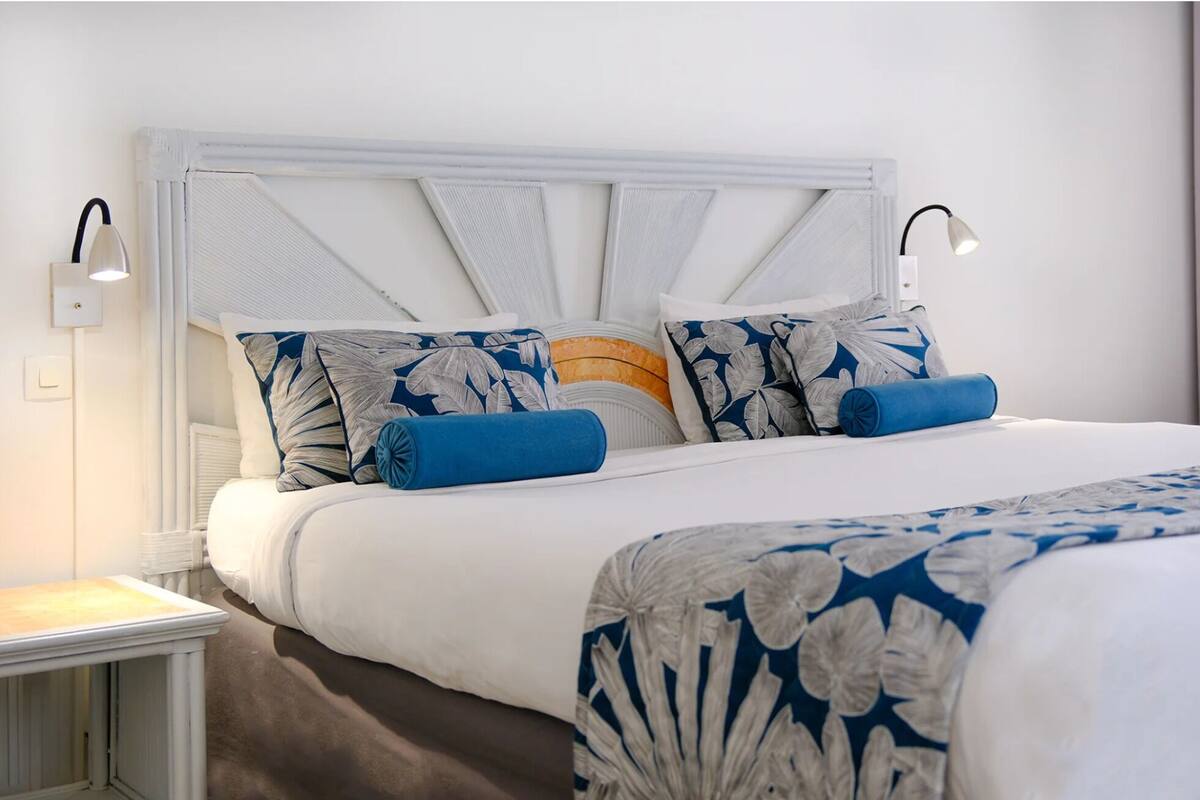 Ein ordentlich bezogenes Bett mit weißem Kopfteil, Kissen mit blau-weißem Muster und einer passenden blau-weißen Tagesdecke. Auf beiden Seiten des Bettes stehen jeweils Nachttischlampen.