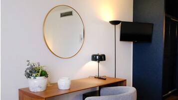 A room with a wooden desk, a grey chair, a round mirror, a small potted plant, a white container, a black floor lamp, and a flat-screen TV mounted on the wall.