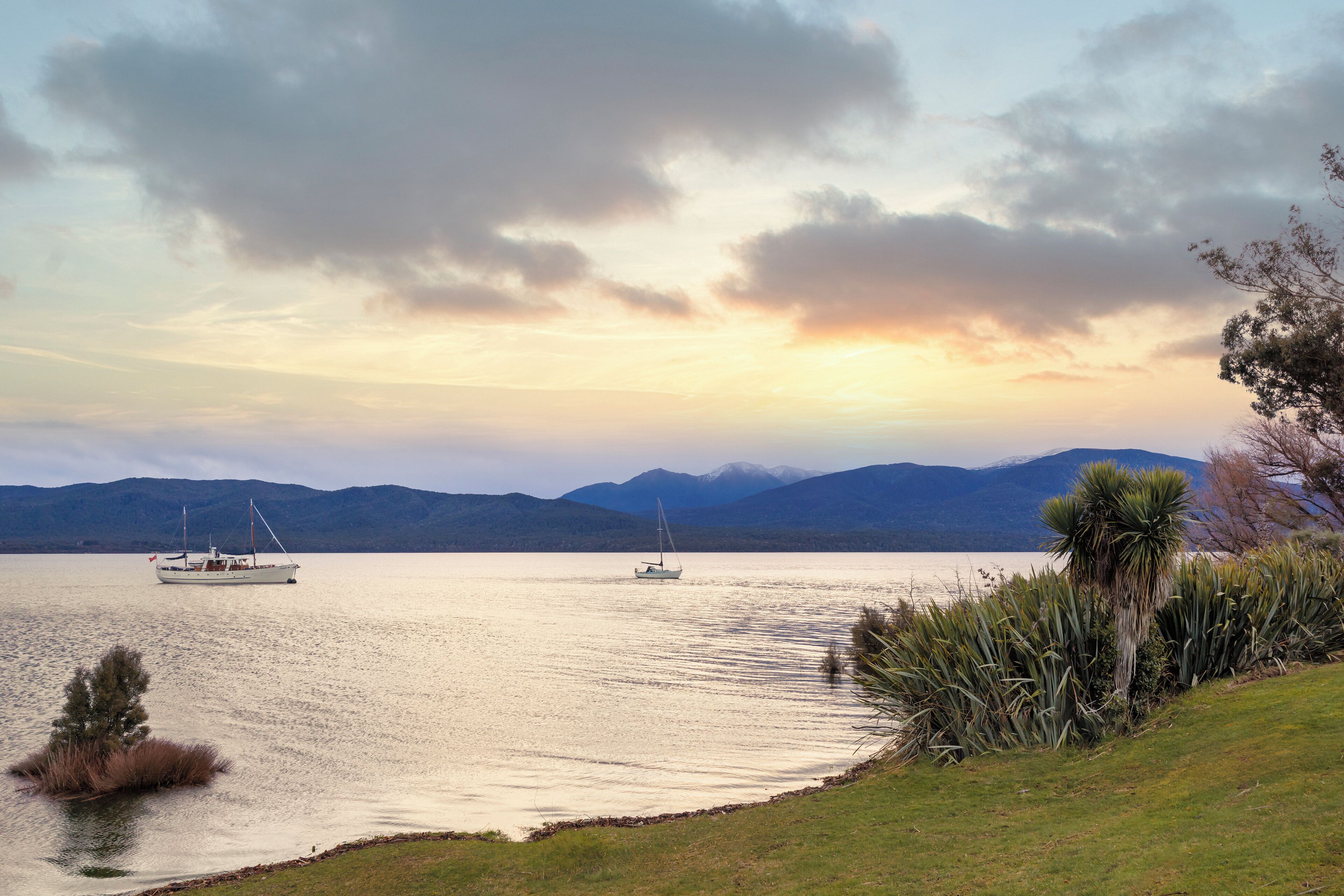 Um lago calmo com barcos, montanhas ao fundo e margens gramadas.
