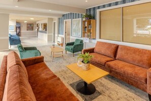 A modern hotel lobby with a seating area featuring brown sofas and green armchairs, a wooden coffee table with a plant, and a reception desk in the background.