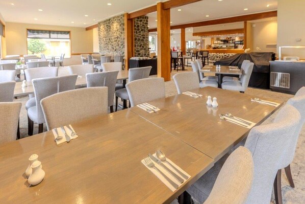 A dining area with a long wooden table set for four, surrounded by grey upholstered chairs. A stone wall and wooden beams are visible in the background.