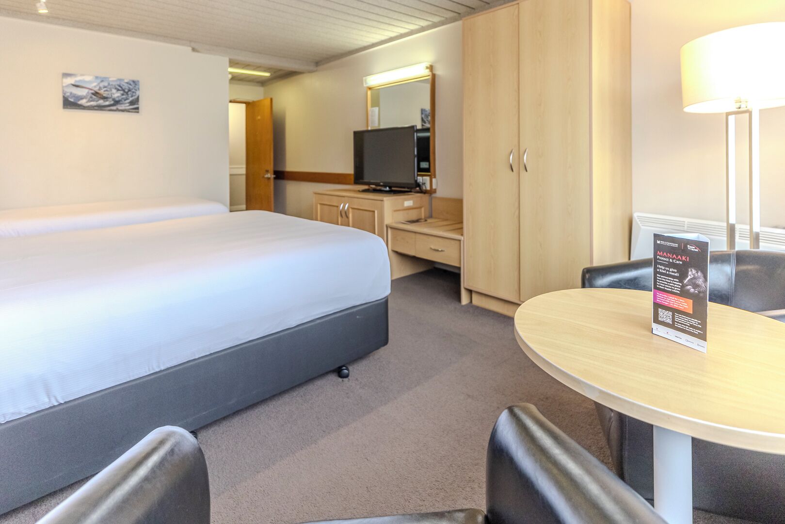 A hotel room with a large bed, a desk with a computer, a chair, and a table with a sign.