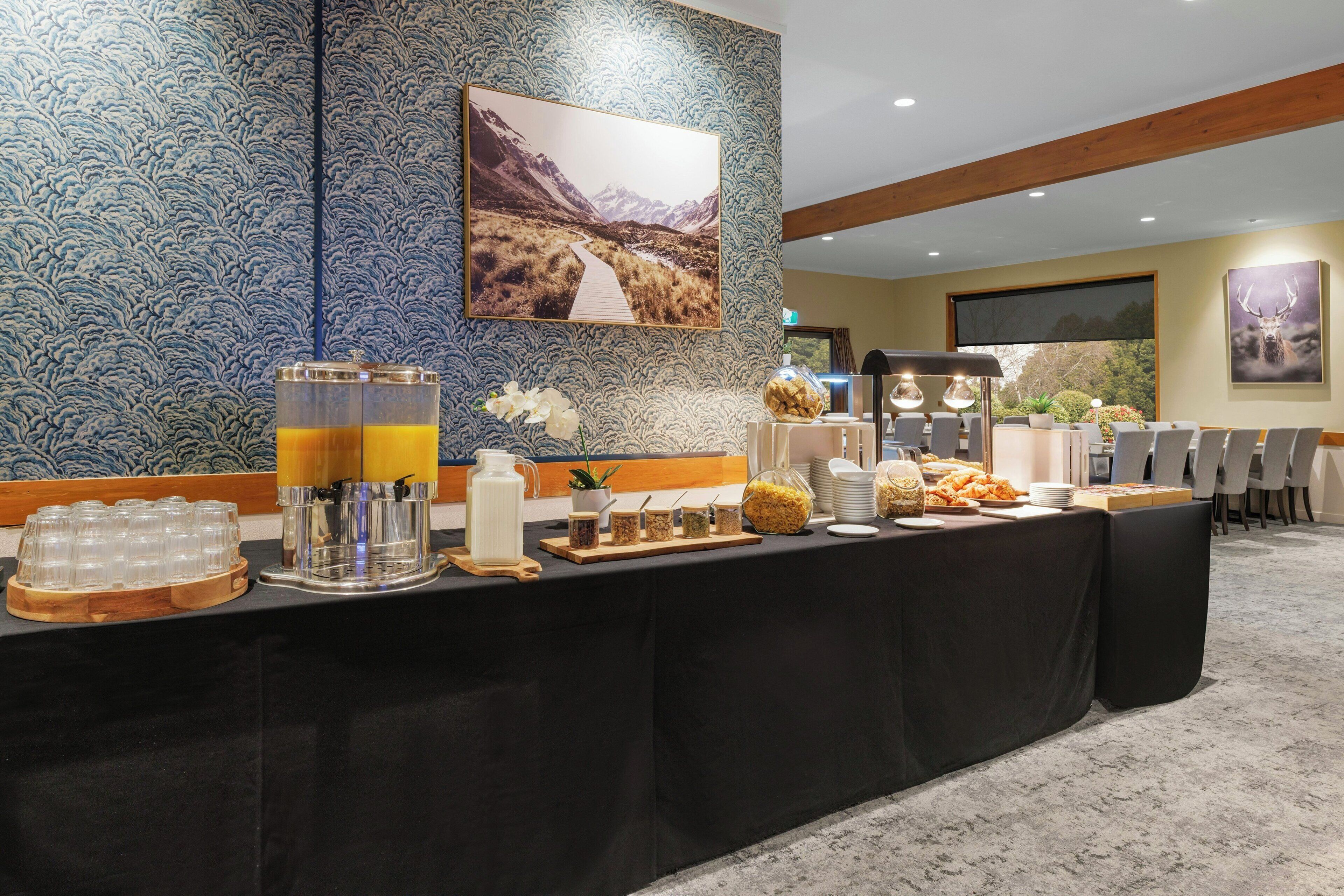 A buffet setup with a variety of food items and beverages, a decorative wall with a mountain landscape, and a seating area with chairs.