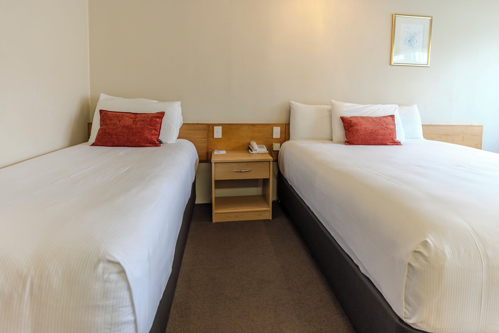 A hotel room with two beds, each with white bedding and red pillows, a wooden nightstand with a phone, and a framed picture on the wall.