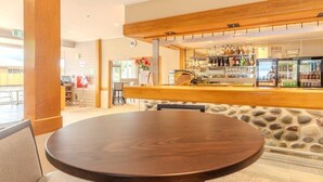 A round wooden table with a cushioned chair in a modern bar area with a stone wall backdrop and a bar counter stocked with various bottles. - Kingsgate Hotel Te Anau (Te Anau)