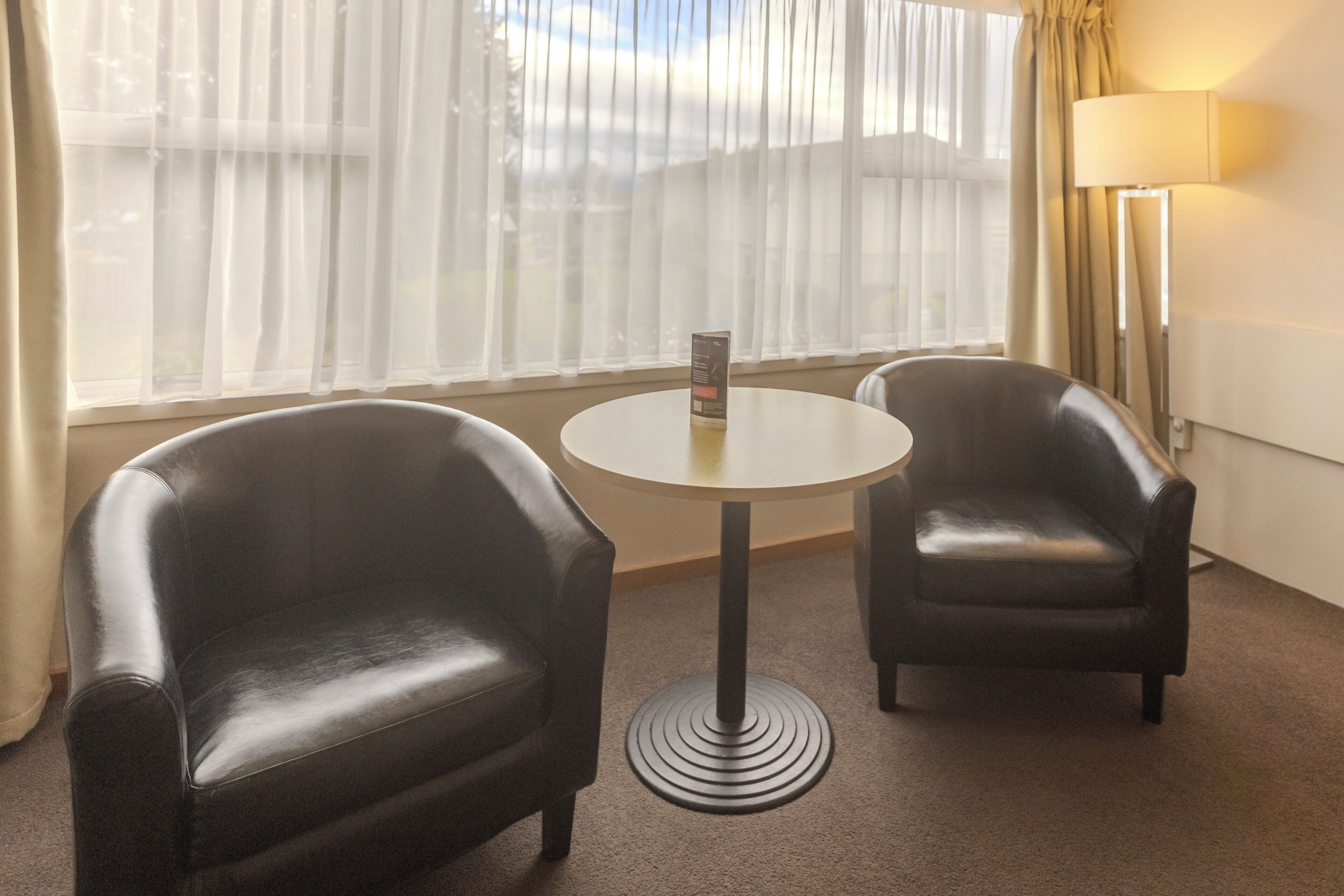A hotel room with two black leather armchairs, a small round table, a standing lamp, and a window with sheer curtains.